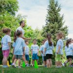 Group of children and teachers playing and learning in a sunny park setting.